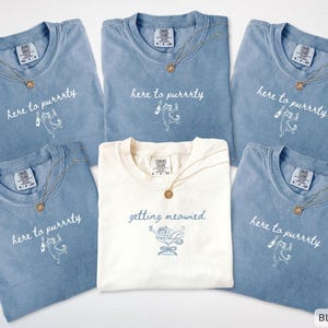 Puede incluir: Se muestran seis camisetas, cinco en azul vaquero y una en marfil. Cada una tiene cuello redondo y un gráfico con el texto "here to purrrty" o "getting meowied" y una ilustración de gato. Una cadena dorada está en cada camiseta.