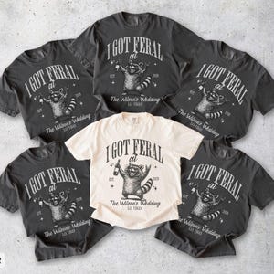 May include: A collection of short-sleeved t-shirts in pepper and ivory. Each shirt features a raccoon graphic with the text "I GOT FERAL at The Wilson's Wedding". The shirts are casual and suitable for a wedding.