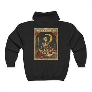 Puede incluir: Sudadera con capucha negra con un diseño de carta del tarot. El gráfico representa un esqueleto meditando con una luna creciente, una vela y las palabras "THE READER" en la parte superior. La palabra "BOOKS" está en la parte inferior de la carta.