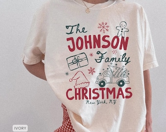May include: An ivory short-sleeve t-shirt featuring red and dark green holiday graphics. The text reads The JOHNSON Family CHRISTMAS New York, NY, accompanied by illustrations of a gingerbread man, a gift, a Santa hat, and a truck.