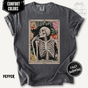May include: Pepper-coloured t-shirt with a tarot card design featuring a skeleton figure, surrounded by flowers, and the text "THE OVERTHINKER." The shirt has rolled-up sleeves and the words "COMFORT COLORS" are printed on the top left.