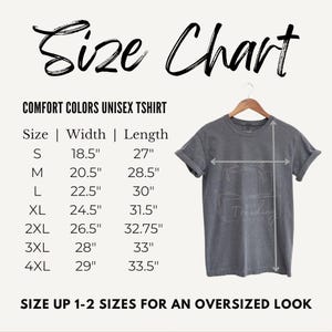 Puede incluir: Una tabla de tallas para una camiseta unisex Comfort Colors, con medidas en pulgadas para las tallas S a 4XL. La tabla incluye las dimensiones de ancho y largo. La imagen tambi&eacute;n muestra una camiseta gris en una percha.
