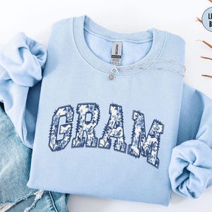 May include: Light blue sweatshirt with the word "GRAM" in a floral design. The sweatshirt is folded with the sleeves tucked in. A silver necklace with a small pendant is on the sweatshirt. The words "LIGHT BLUE" are in a circle in the upper right corner.