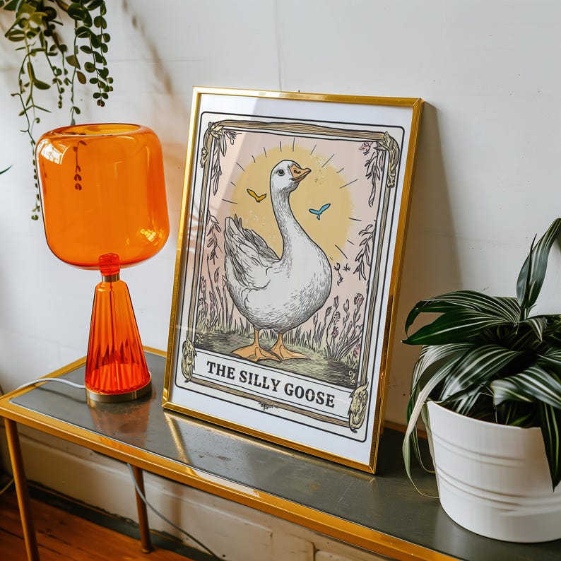 May include: A framed print with a gold frame and a white background. The print features a cartoon illustration of a white goose with a pink beak and blue eyes. The goose is standing in a field of green grass and flowers. The text "The Silly Goose" is printed below the goose.