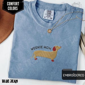 May include: Blue jean colored t-shirt with an embroidered dachshund design and the text "WEENIE MOM". The shirt has a crew neck and short sleeves. A gold necklace with a pendant is on the shirt. The shirt is labeled "COMFORT COLORS".