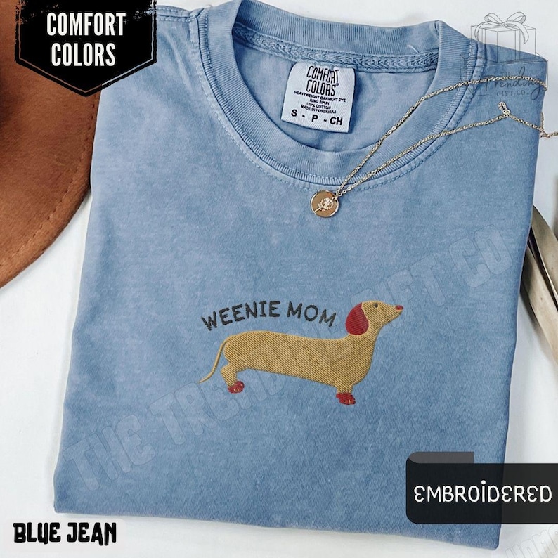 May include: Blue jean colored t-shirt with an embroidered dachshund design and the text "WEENIE MOM". The shirt has a crew neck and short sleeves. A gold necklace with a pendant is on the shirt. The shirt is labeled "COMFORT COLORS".