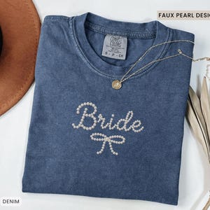 Puede incluir: Una camiseta azul denim con la palabra "Bride" escrita con perlas de imitación y un lazo de perlas. Un collar de oro con un colgante descansa sobre la camisa. La camiseta tiene una etiqueta Comfort Colors. Las palabras "Faux Pearl Design" están impresas en la parte superior derecha.