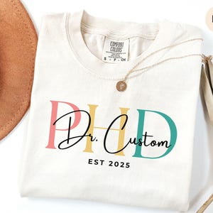 May include: Ivory t-shirt with the text "PhD Dr. Custom EST 2025" in pink, yellow, and teal lettering.