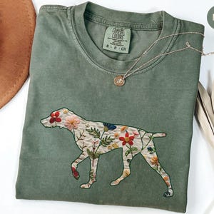 Puede incluir: Una camiseta verde musgo con un diseño floral bordado de un perro. La camiseta tiene el texto "COMFORT COLORS" y "S - P - CH" en la etiqueta.