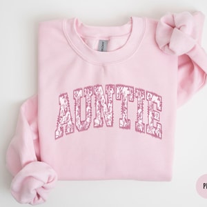 以下が含まれることがあります： 花柄のデザインで「AUNTIE」と書かれたピンク色のスウェットシャツです。