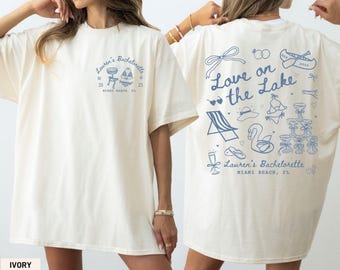 May include: Ivory-colored t-shirt with blue text and graphics. The front features "Lauren's Bachelorette" and a graphic of a bar and beach chair. The back says "Love on the Lake" with illustrations of a beach chair, swan, and champagne glasses.
