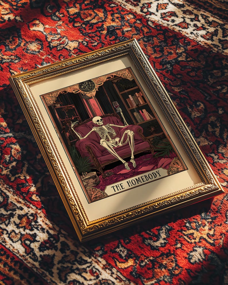 May include: Framed artwork featuring a skeleton lounging in a burgundy armchair, holding a wine glass. The artwork is set in a room with bookshelves and a patterned rug. The text "THE HOMEBODY" is at the bottom.