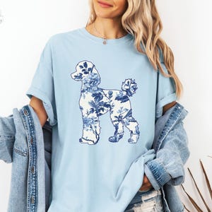 May include: A light blue t-shirt with a blue and white floral poodle silhouette design. The shirt is made of a soft, comfortable chambray fabric.