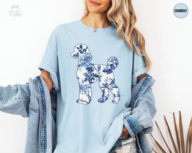May include: A light blue t-shirt with a blue and white floral poodle silhouette design. The shirt is made of a soft, comfortable chambray fabric.