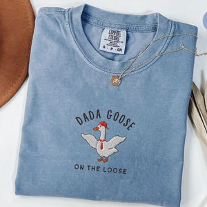 May include: A blue jean t-shirt with the text "Dada Goose On The Loose" and an embroidered image of a goose wearing a red hat.