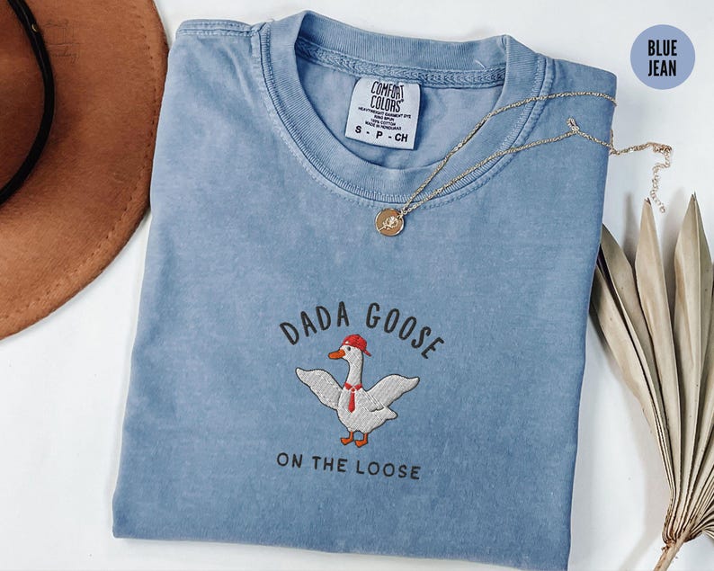 May include: A blue jean t-shirt with the text "Dada Goose On The Loose" and an embroidered image of a goose wearing a red hat.