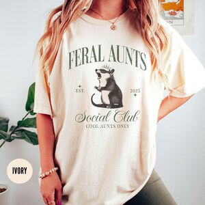 May include: Ivory t-shirt featuring a graphic of a possum wearing a crown and the text "Feral Aunts Social Club Cool Aunts Only" with the text "Est. 2025" below the graphic.