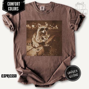 May include: Espresso-coloured t-shirt with a sepia-toned graphic of a raccoon wearing sunglasses and holding a martini. The shirt has the text "COMFORT COLORS" and "UNISEX SIZING" on it.