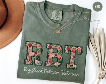 May include: A moss green t-shirt featuring embroidered letters "RBT" filled with strawberries and flowers. The words "Registered Behavior Technician" are written below. A gold necklace is draped across the shirt. The shirt is a size S.