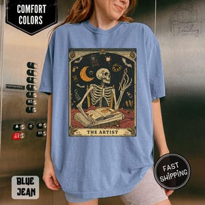 Puede incluir: Camiseta azul claro con un dise&ntilde;o de carta del tarot que presenta un artista esqueleto, una luna creciente y materiales de pintura. El texto "THE ARTIST" est&aacute; en la parte inferior de la carta. La camiseta tiene las palabras "COMFORT COLORS" y "BLUE JEAN".