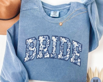 May include: A blue jean sweatshirt with the word "BRIDE" in white letters with a floral pattern. The sweatshirt has a distressed look.