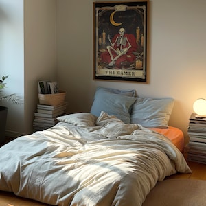 Puede incluir: Una cama beige con un edred&oacute;n blanco y una almohada azul. Un p&oacute;ster enmarcado con un esqueleto sentado en un trono y el texto "The Gamer" est&aacute; colgado en la pared detr&aacute;s de la cama.