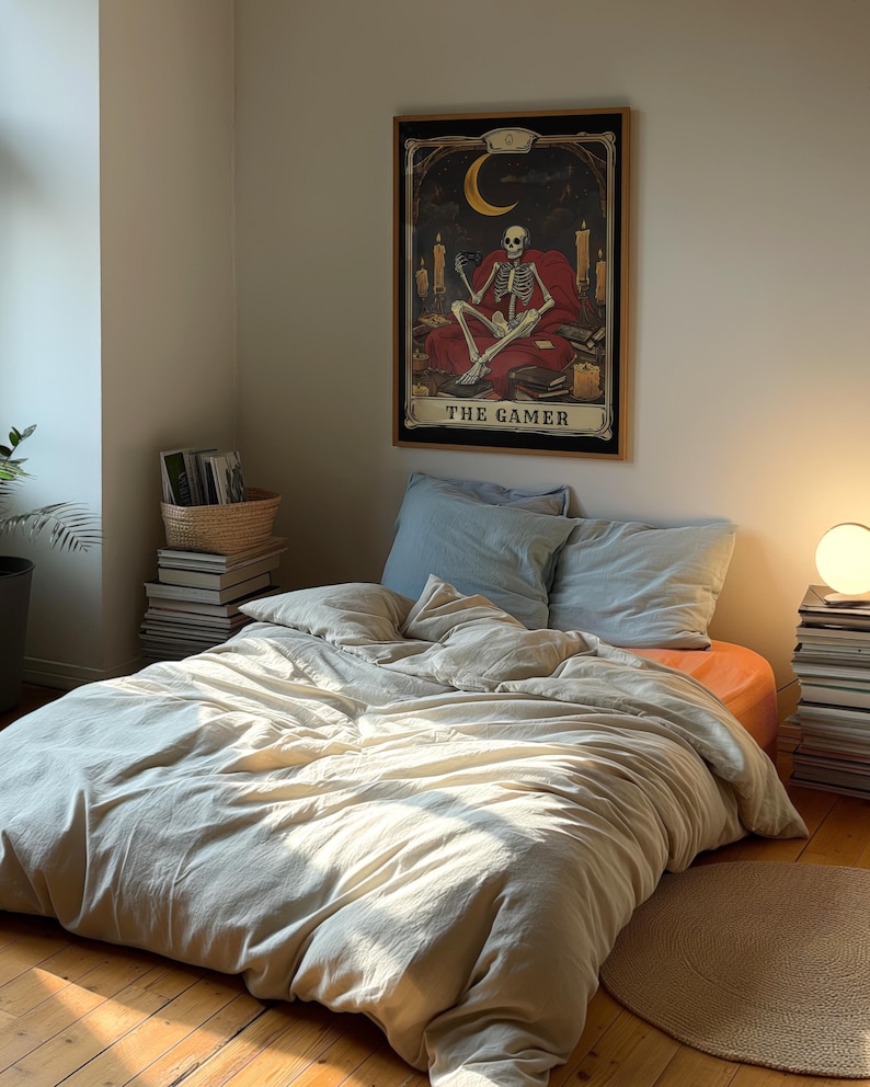 Puede incluir: Una cama beige con un edred&oacute;n blanco y una almohada azul. Un p&oacute;ster enmarcado con un esqueleto sentado en un trono y el texto "The Gamer" est&aacute; colgado en la pared detr&aacute;s de la cama.