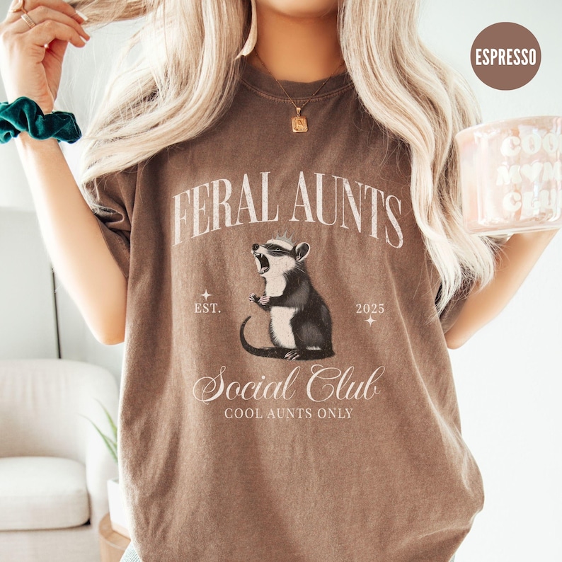 May include: A brown t-shirt with the text "Feral Aunts Social Club Cool Aunts Only" and a cartoon illustration of a rodent. The text "Est. 2025" is also on the shirt.
