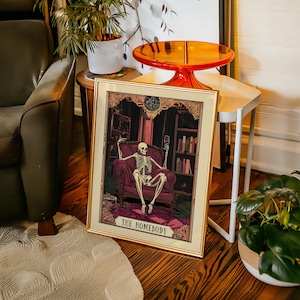 May include: Framed art print featuring a skeleton lounging in a plush armchair, holding a wine glass. The artwork has a gothic theme with a dark color palette and the text "THE HOMEBODY" at the bottom. The frame is gold.