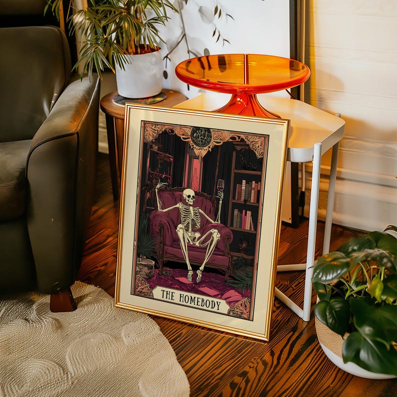 May include: Framed art print featuring a skeleton lounging in a plush armchair, holding a wine glass. The artwork has a gothic theme with a dark color palette and the text "THE HOMEBODY" at the bottom. The frame is gold.