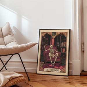 May include: Framed art print featuring a skeleton seated in a red armchair, holding a wine glass. The artwork has a dark, gothic theme with bookshelves and the text "THE HOMEBODY" at the bottom. The frame is black.