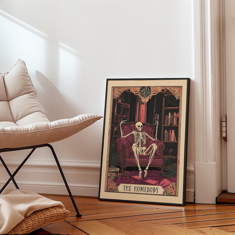 May include: Framed art print featuring a skeleton seated in a red armchair, holding a wine glass. The artwork has a dark, gothic theme with bookshelves and the text "THE HOMEBODY" at the bottom. The frame is black.