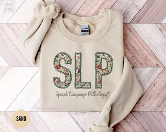 May include: A beige sweatshirt with the words "SLP Speech Language Pathologist" printed on the front. The letters are made of a floral pattern.