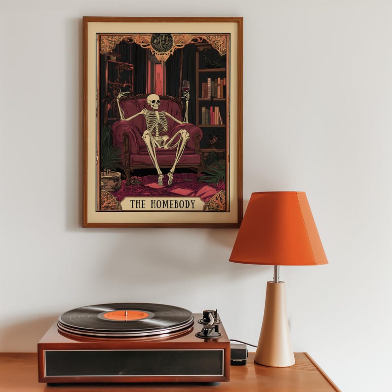 May include: A framed art print featuring a skeleton in a burgundy armchair, holding a wine glass. The print is labeled "THE HOMEBODY." A vintage record player and orange lamp are in the foreground.
