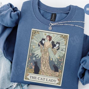 May include: Indigo blue sweatshirt with a tarot card design featuring a woman and black and white cats. The card reads "The Cat Lady." A silver necklace with a sun pendant is draped across the sweatshirt.