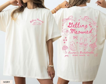 May include: Ivory-colored t-shirt with the text "Getting Meowied" in pink on the front and back. The back design includes cat illustrations, bows, champagne glasses, and the text "Lauren's Bachelorette, Miami Beach, FL."