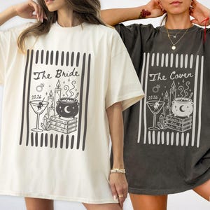 May include: An ivory t-shirt and a pepper gray t-shirt, each featuring a black graphic print of a martini glass, a cauldron, candles, crystals, and a stack of books. The ivory shirt reads The Bride 2026, while the pepper shirt reads The Coven 2026.