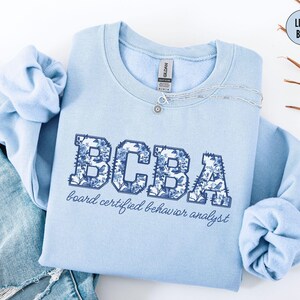 May include: Light blue sweatshirt with the letters "BCBA" in a floral design. The words "board certified behavior analyst" are printed below the letters. A silver necklace with a pendant is on the sweatshirt.