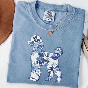 May include: A blue jean t-shirt with a white silhouette of a poodle with a floral pattern. The shirt has a tag that says "Comfort Colors" and "S - P - CH".