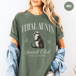 May include: A green t-shirt with the text "Feral Aunts Social Club Cool Aunts Only" and a cartoon image of a possum wearing a crown. The text "Est. 2025" is also on the shirt.