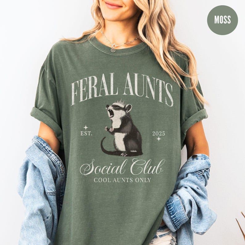 May include: A green t-shirt with the text "Feral Aunts Social Club Cool Aunts Only" and a cartoon image of a possum wearing a crown. The text "Est. 2025" is also on the shirt.