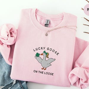 May include: Pink sweatshirt with a graphic of a goose holding shamrocks and the text "Lucky Goose on the Loose".