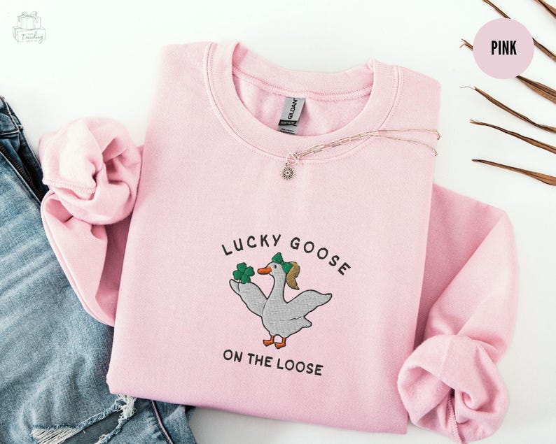 May include: Pink sweatshirt with a graphic of a goose holding shamrocks and the text "Lucky Goose on the Loose".