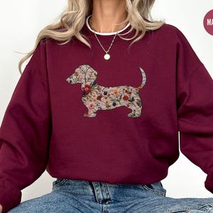 Może przedstawiać: Bordowa bluza z nadrukiem w kształcie sylwetki psa rasy dachshund ozdobionej kwiatami. Bluza ma okrągły dekolt i długie rękawy.