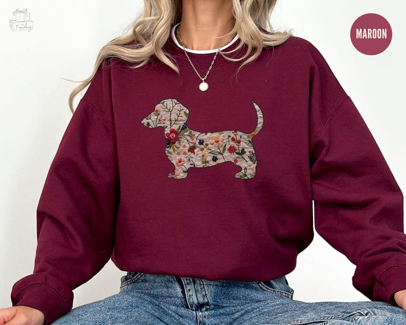 Może przedstawiać: Bordowa bluza z nadrukiem w kształcie sylwetki psa rasy dachshund ozdobionej kwiatami. Bluza ma okrągły dekolt i długie rękawy.