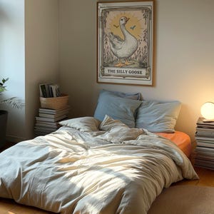 May include: A framed print of a goose with the text "The Silly Goose" in a bedroom setting. The print is on a wall above a bed with a white duvet and blue pillows.