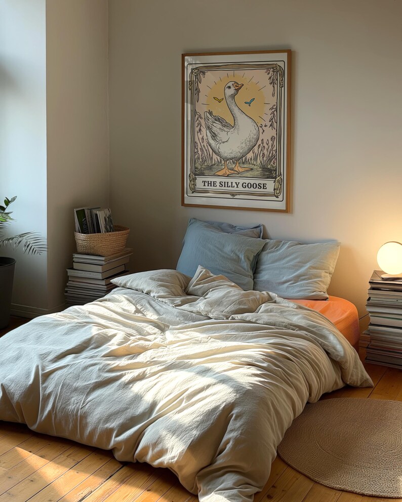 May include: A framed print of a goose with the text "The Silly Goose" in a bedroom setting. The print is on a wall above a bed with a white duvet and blue pillows.
