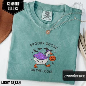 May include: Light green t-shirt with the embroidered phrase "SPOOKY GOOSE ON THE LOOSE". The design features a goose wearing a witch hat and cape, carrying a jack-o'-lantern. The shirt has a Comfort Colors label.