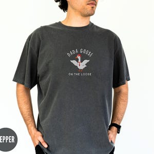 May include: A dark gray t-shirt with a white goose wearing a red hat and a red tie. The text "Dada Goose On The Loose" is printed on the shirt.