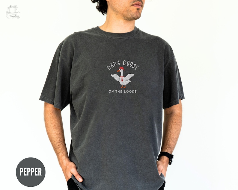 May include: A dark gray t-shirt with a white goose wearing a red hat and a red tie. The text "Dada Goose On The Loose" is printed on the shirt.